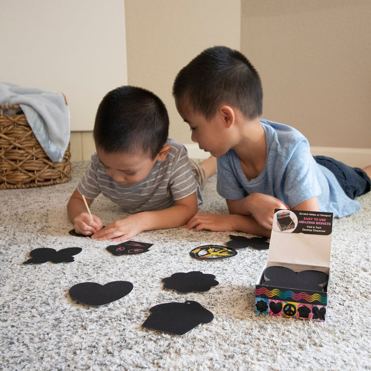 A kid playing with The Melissa & Doug Scratch Art® Box of 125 Friendship-Themed Shaped Notes in Desktop Dispenser (Approx. 3.5” x 3.5” Each Note)