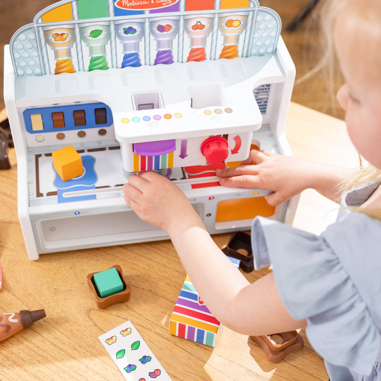 A kid playing with The Melissa & Doug Wooden Chocolate Factory Pretend Play Set, Play Food Candy Maker for Boys and Girls