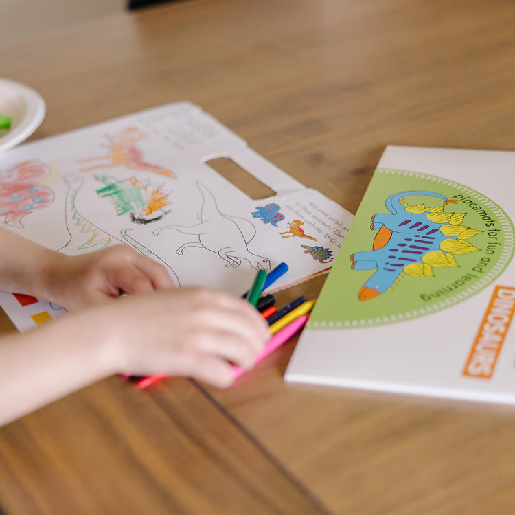 A kid playing with The Melissa & Doug Playmats Dinosaurs Take-Along Paper Coloring And Learning Activity Pads (24 Pages)