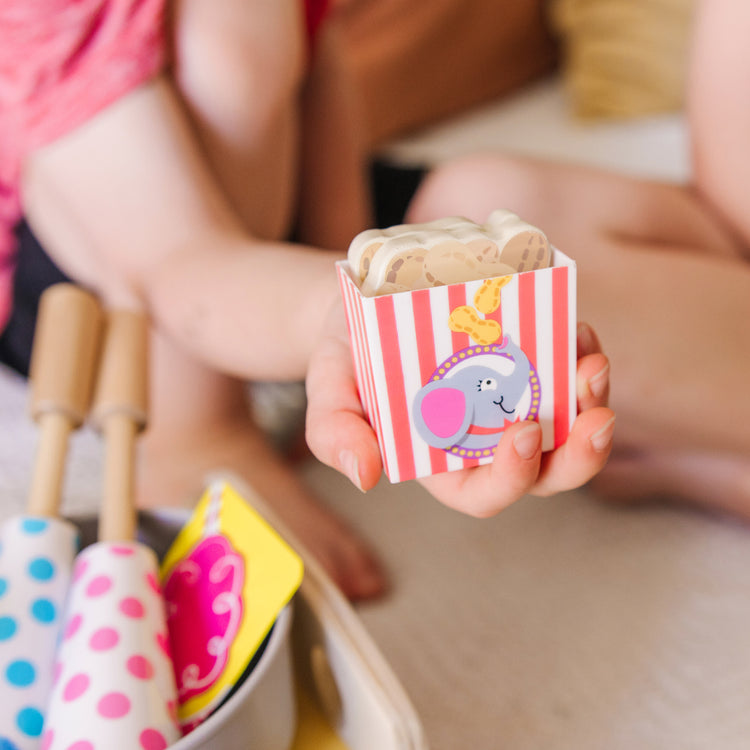 A kid playing with The Melissa & Doug Fun at the Fair! Wooden Carnival Candy Tabletop Cart and Play Food Set