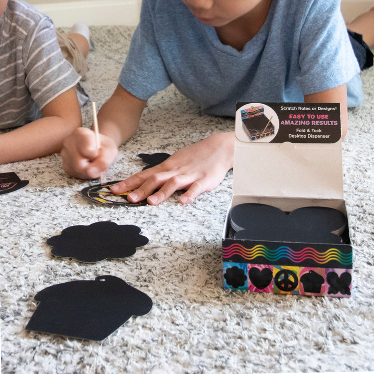 A kid playing with The Melissa & Doug Scratch Art® Box of 125 Friendship-Themed Shaped Notes in Desktop Dispenser (Approx. 3.5” x 3.5” Each Note)