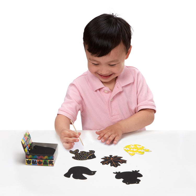 A child on white background with The Melissa & Doug Scratch Art® Box of 125 Nature-Themed Shaped Notes in Desktop Dispenser (Approx. 3.5” x 3.5” Each Note)