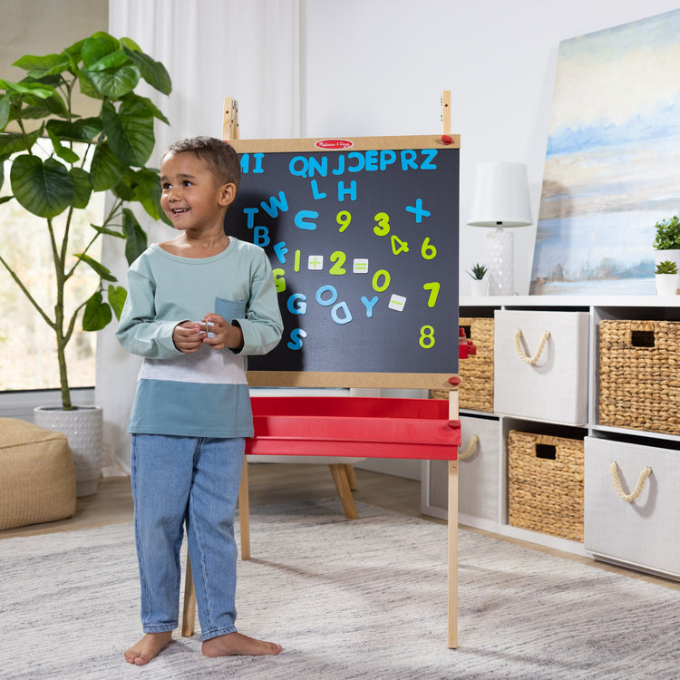 A kid playing with The Melissa & Doug Deluxe Magnetic Standing Art Easel With Chalkboard, Dry-Erase Board, and 39 Letter and Number Magnets