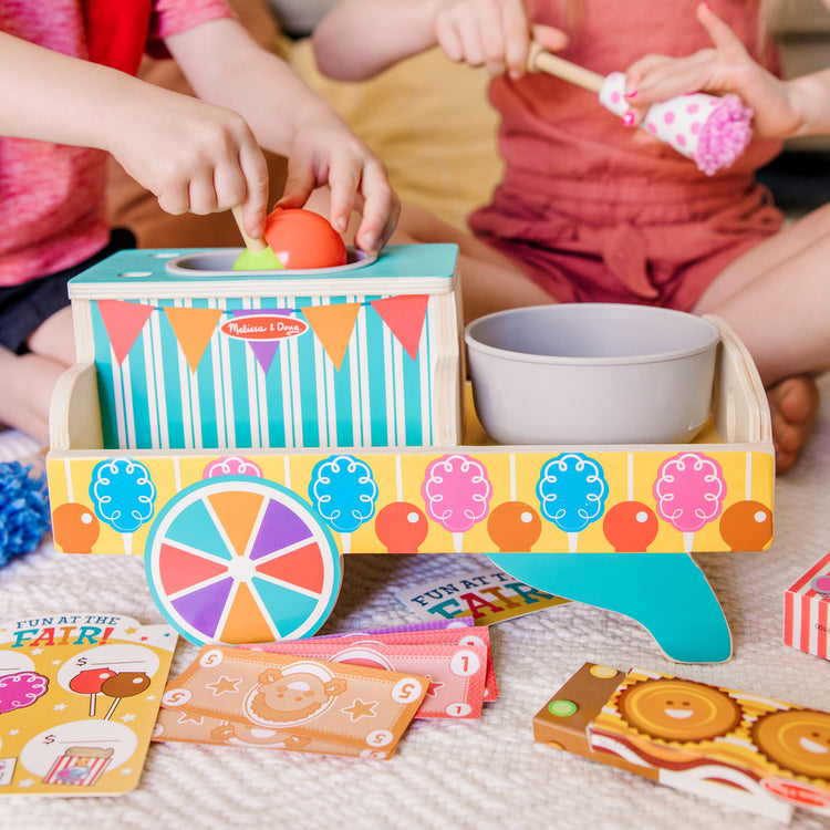 A kid playing with The Melissa & Doug Fun at the Fair! Wooden Carnival Candy Tabletop Cart and Play Food Set