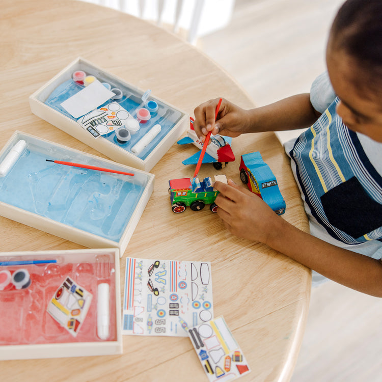 A kid playing with The Melissa & Doug Decorate-Your-Own Wooden Craft Kits 3-Pack - Plane, Train, and Race Car