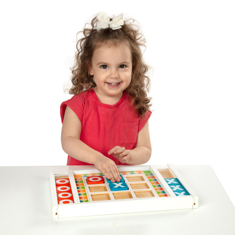 A child on white background with The Melissa & Doug Wooden Tic-Tac-Toe Board Game with 10 Self-Storing Wooden Game Pieces (12.5” W x 8.5” L x 1.25” D)